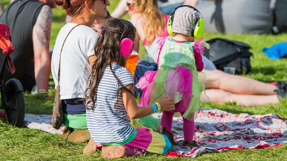 Two young children wearing ear defenders as they relax on a blanket next to a female.