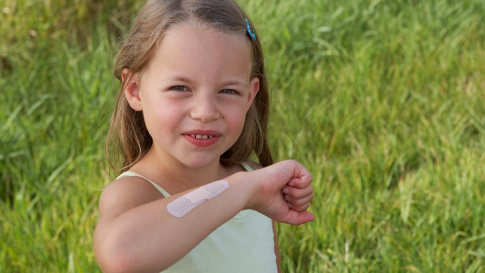 A young girl with her right arm raised in front of her showing that she has a plaster on her arm.