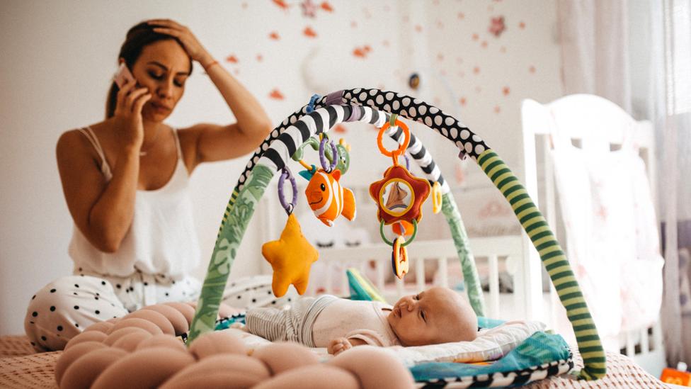 Mother on the phone looking worried cross legged on bed. Baby lying on play mat on bed.