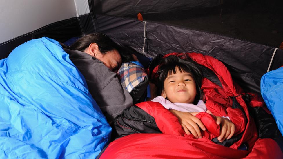 A grownup asleep in a blue sleeping bag who is lay next to a young child, who is lay awake and smiling at the camera, in a red sleeping bag.