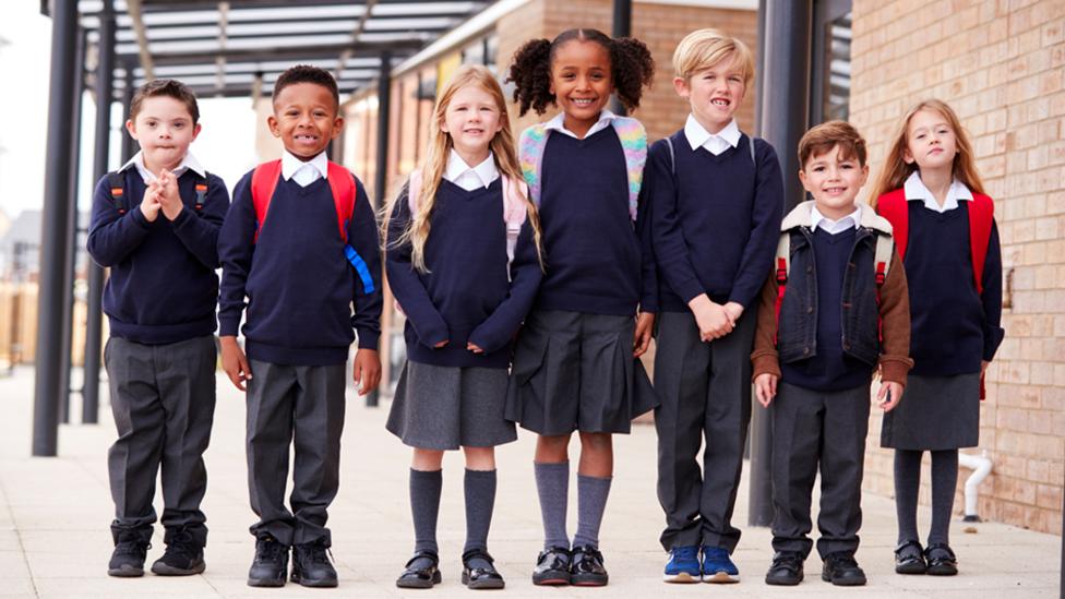 Seven young children all wearing a school uniform stand in a line while smiling.