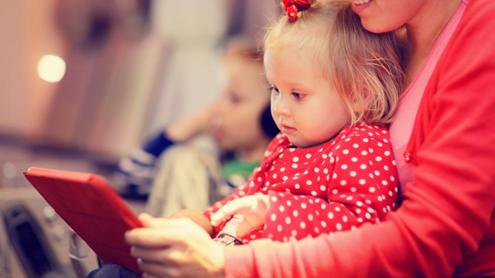 Young child and her mother sat on a plane playing on a tablet.