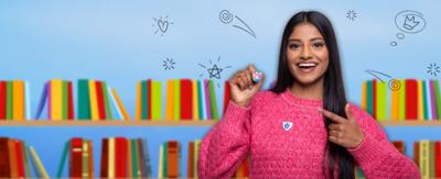 Blue Peter presenter Shini is wearing a pink jumper and holding the Book badge. She is pointing at it and smiling.