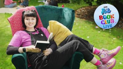 Blue Peter presenter, Abby, lies across an armchair in a green garden holding a book and smiling. She wears a pink t shirt, pink trainers and black dungarees.