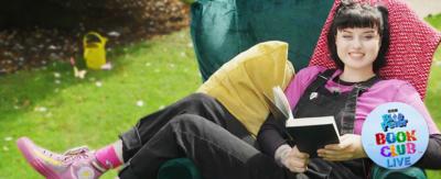 Blue Peter presenter, Abby, lies across an armchair in a green garden holding a book and smiling. She wears a pink t shirt, pink trainers and black dungarees.