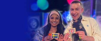 Blue Peter presenter Shini is smiling whilst holding an iced biscuit with the Swedish flag on it. Next to her stands Olly Alexander, who holds another biscuit but with the Union Jack flag on it.
