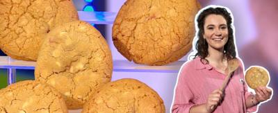 Great British Bake Off contestant Tasha Stone is smiling in a pink shirt whilst holding a wooden spoon in one hand and a white chocolate chip cookie in the other.