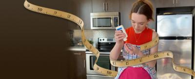 A young girl is in her kitchen, she is holding a pot of glue and is wrapped in measuring tape.