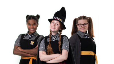 Three young girls in school uniforms, capes and pointed witch hats all smiling to the camera. Above them is a sign that says "The Worst Witch"