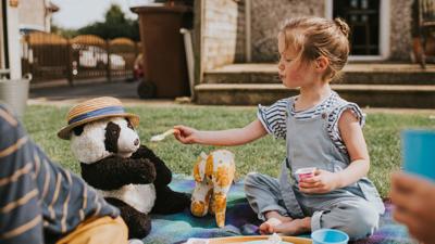 Young girl is sitting outside, holding a spoon to a toy pandas mouth.