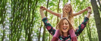 Mother has daughter sitting on her shoulders, they are holding hands and smiling. Background is trees and forest.