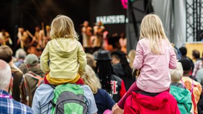 Two young children sat on grownups shoulders. Both are wearing bright colours. They are stood in a crowd watching a performance on a large stage.
