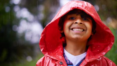 Young boy wearing a red raincoat with his hood up, smiling into camera while it is raining.