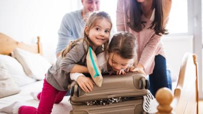 Two young girls helping parents shut a full and slightly overflowing suitcase on top of a bed.