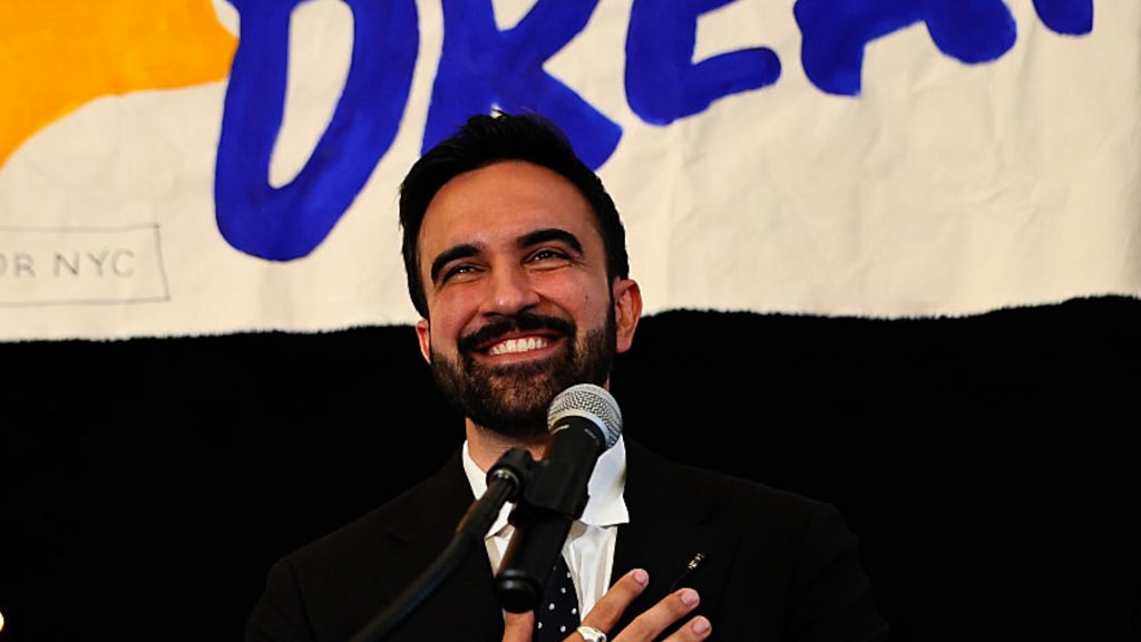 New York mayoral candidate, State Rep. Zohran Mamdani in front of a mic with hand on his heart.