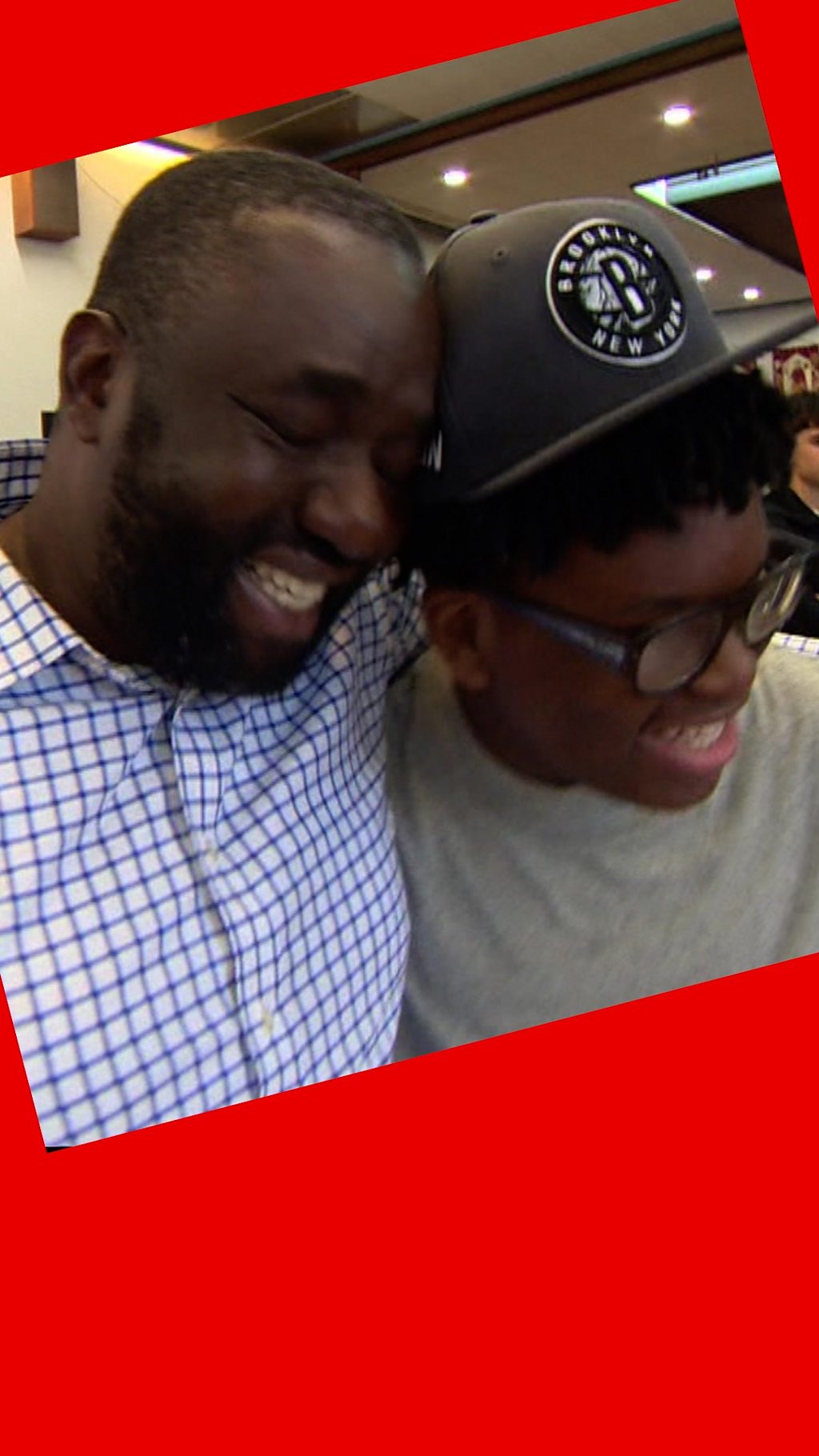 Two men embrace and smile - one is wearing a baseball cap