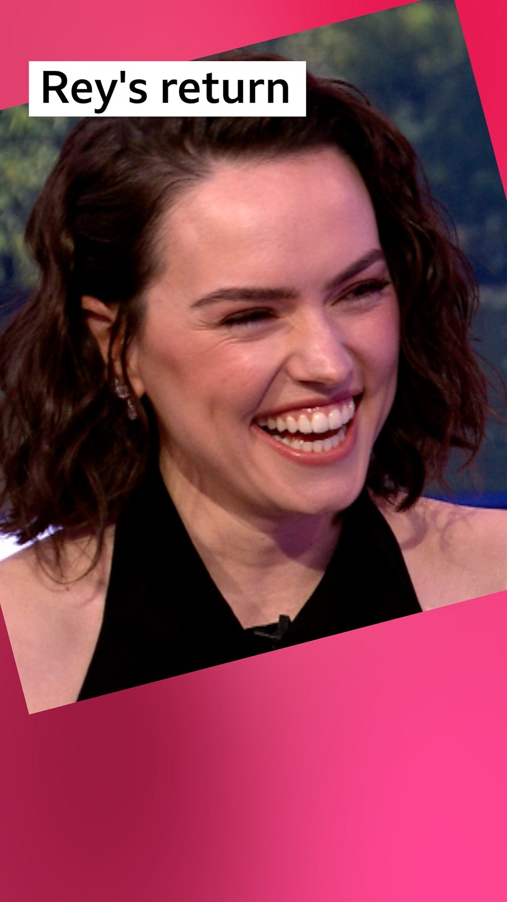 A woman, Daisy Ridley, smiles - she has shoulder length wavy brown hair