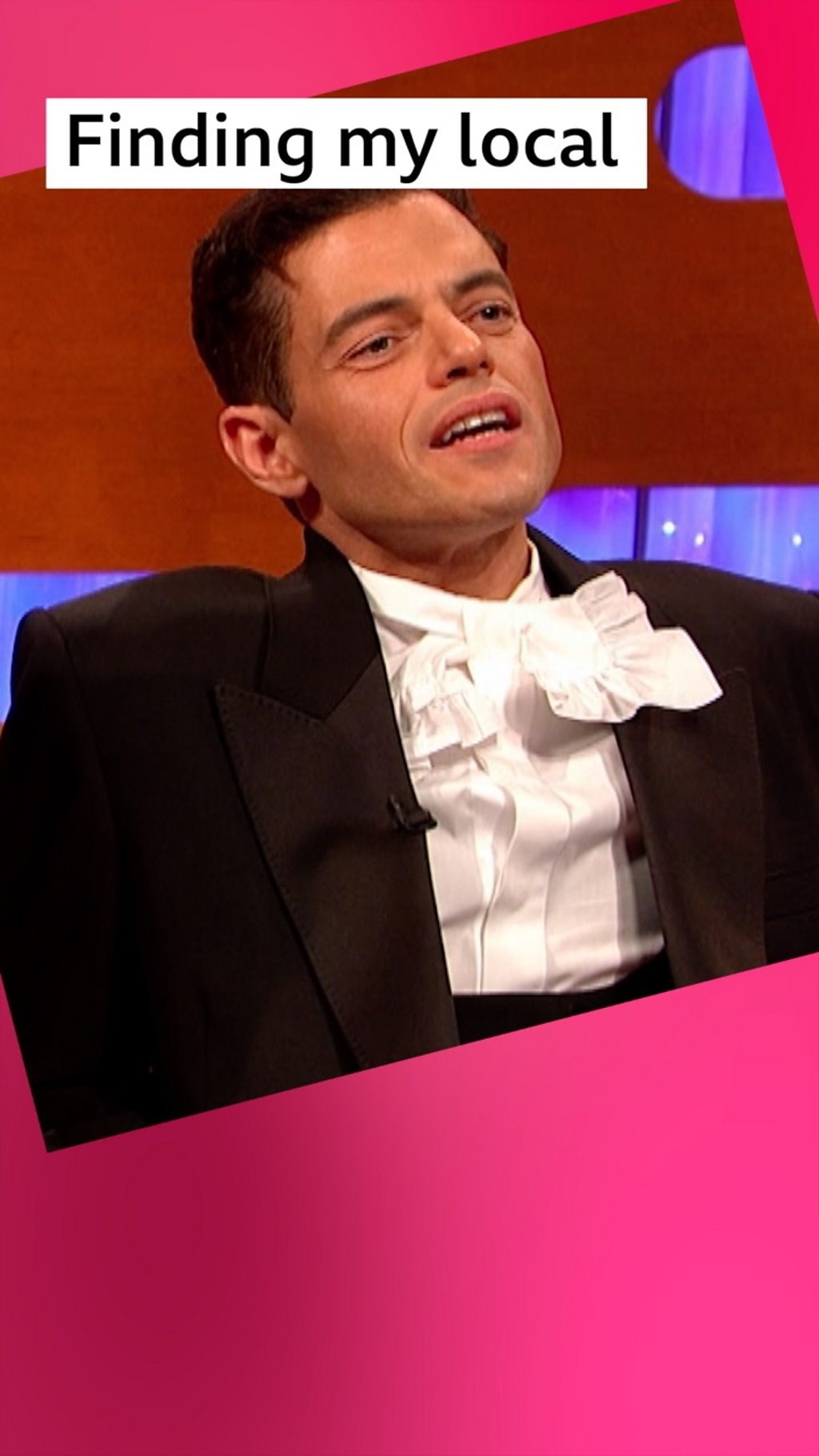 A man, Rami Malek, sits on a sofa - he has short dark hair and wears a white ruffled shirt and a black jacket