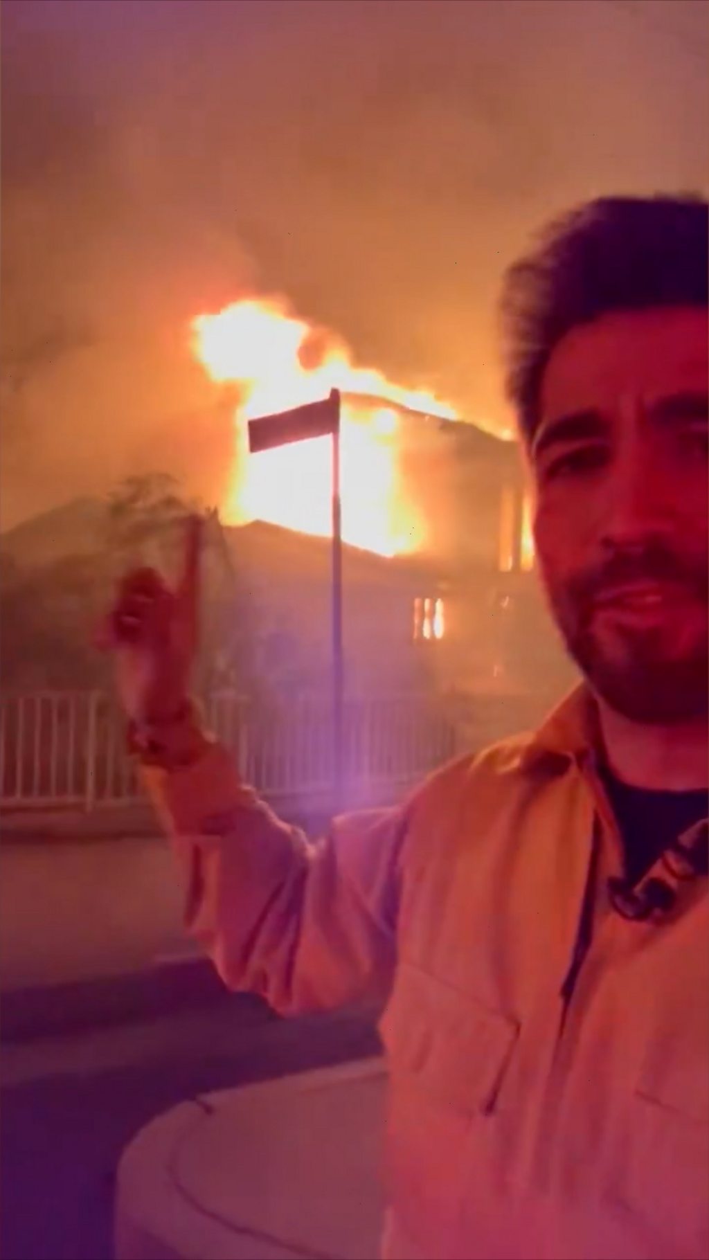 A man stands taking a selfie-style video pointing to a house behind him in flames