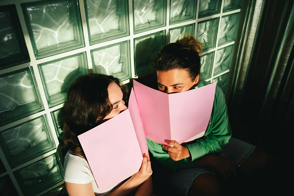 Two friends smile at each other from behind pink menus (Credit: Getty Images)