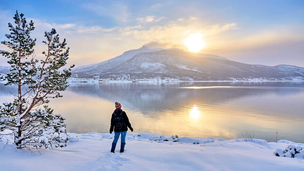 Person standing on a snowy shoreline overlooking a calm lake at sunrise with snow-covered mountain reflected in water (Credit: Alamy)