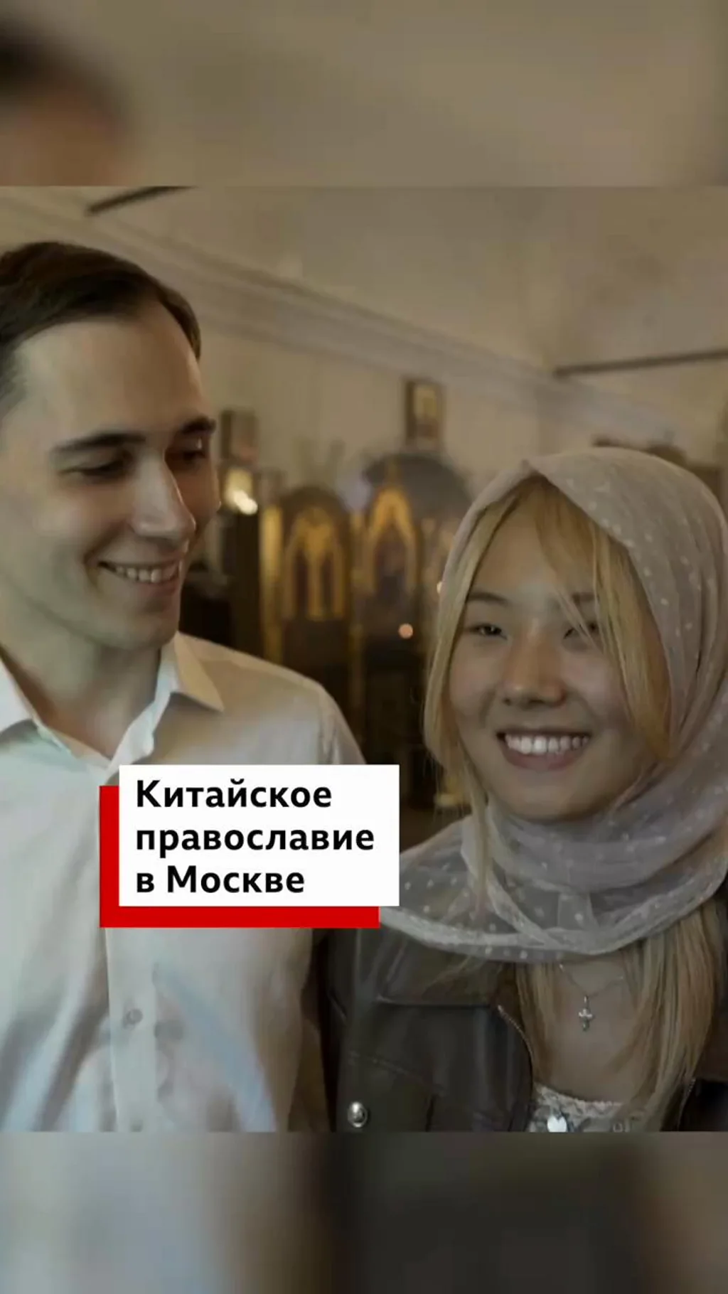 Молодой русский мужчина и женщина-китаянка в платке с улыбкой смотрят друг на друга в православной церкви