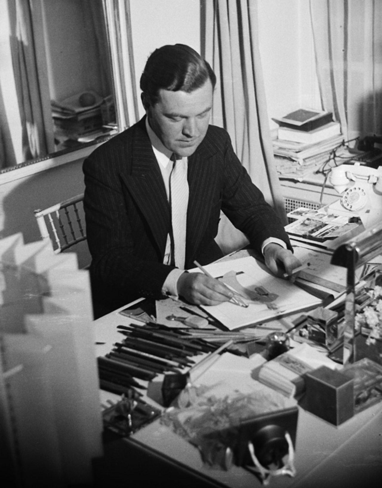 Norman Hartnell, an important designer, designing clothes at his desk.
