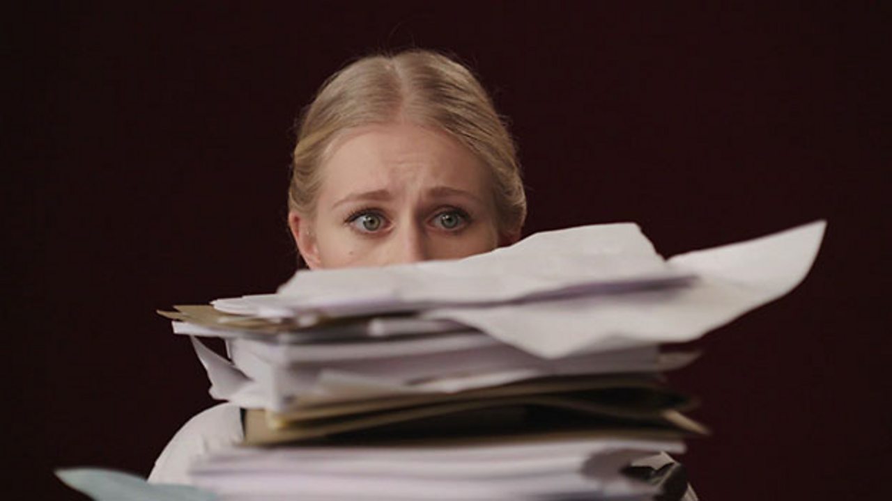 Girl's face is nearly covered by the large pile of work that she is sat behind