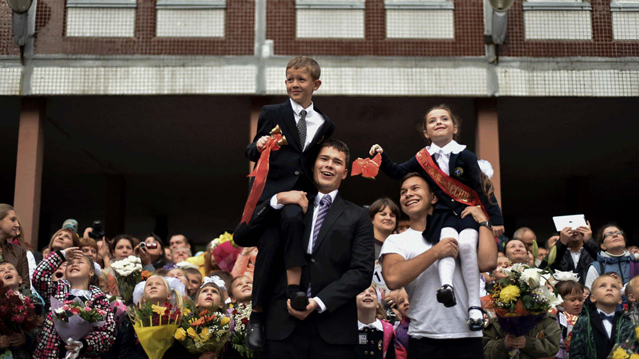 Two young students sit on older students' shoulders, ringing bells