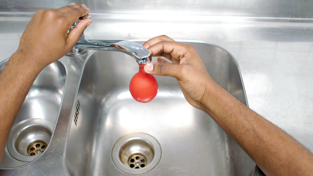 The two hands then fill the red balloon with water over the sink.