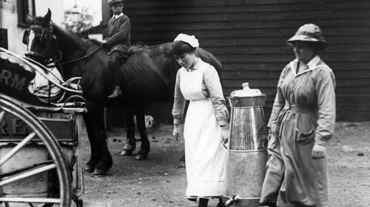 Two milkmaids loading a large milk can onto a horse and cart in the early 1900s