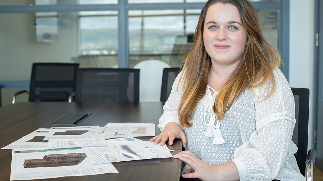 a young woman sits at a desk in an office with papers in front of her