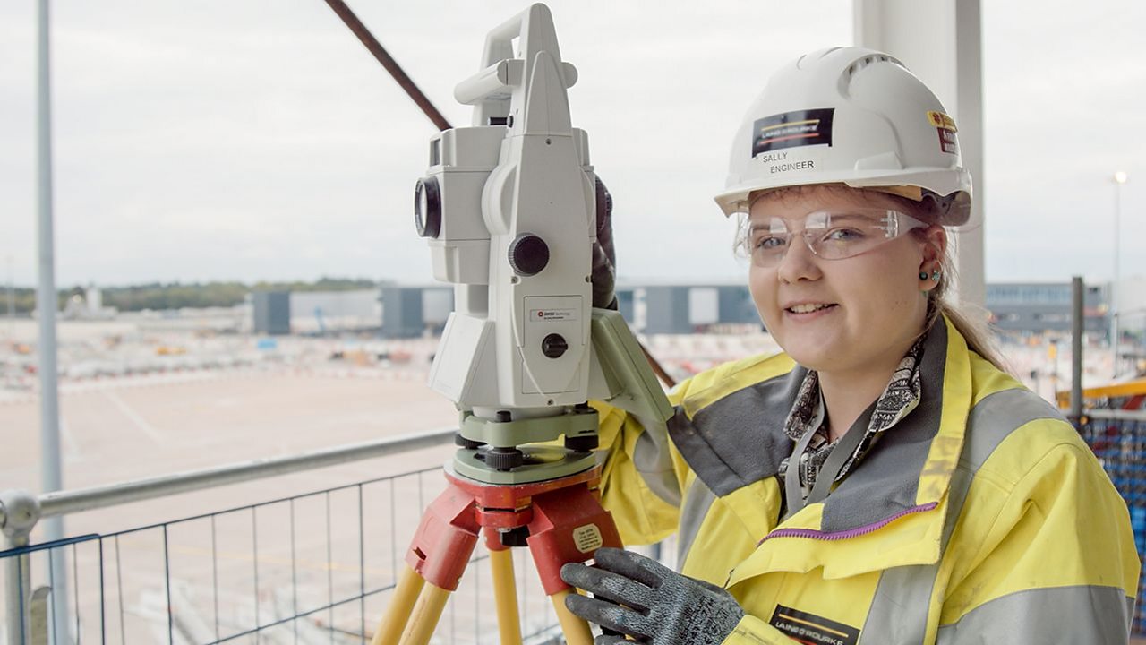 Sally wears a hard hat and operates a surveying machine on a construction site
