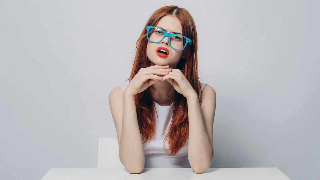 A redhead with with glasses stares from across the table as if interrogating the viewer.