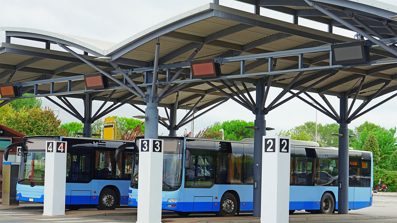 A photo of a bus station. There are three buses parked up at bus stops.