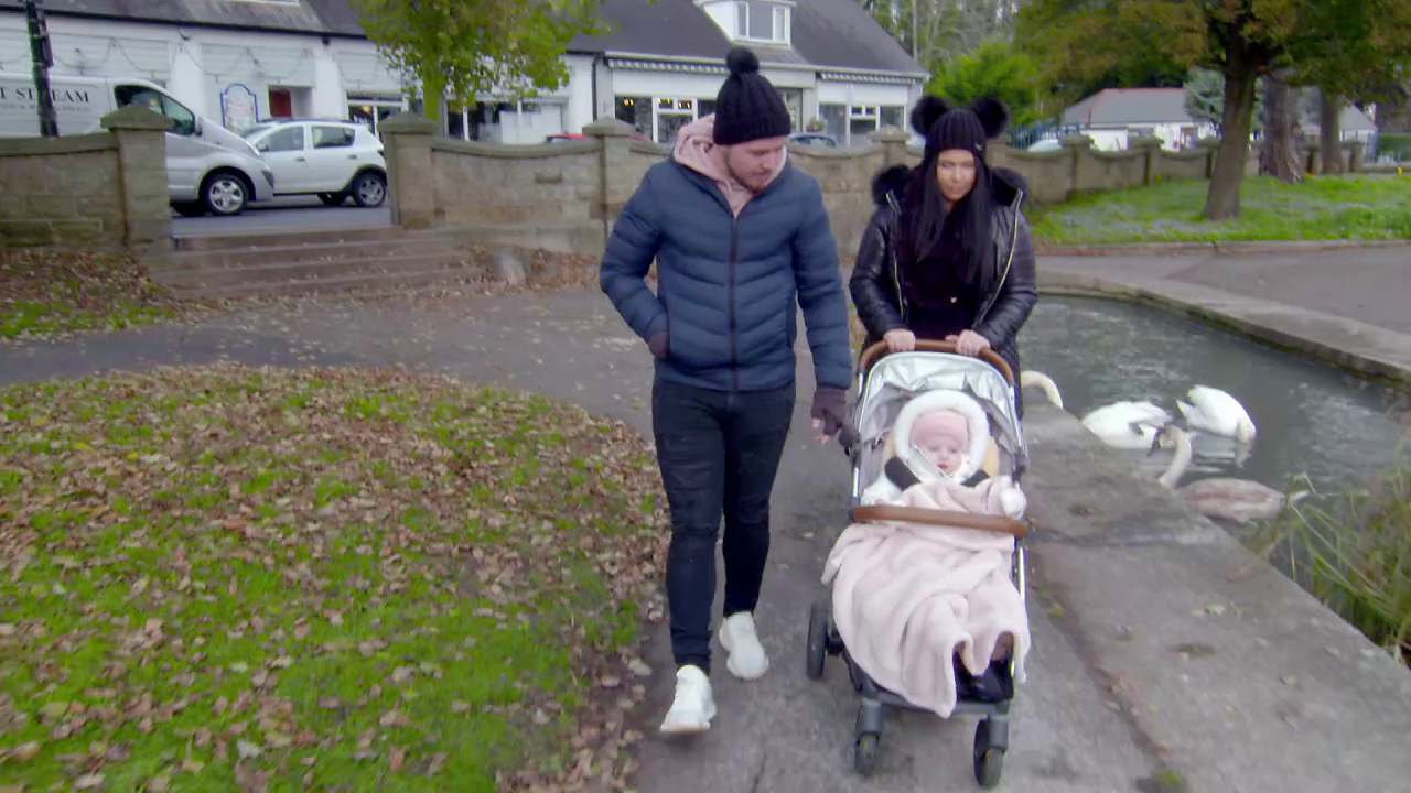 Man and woman in park wearing bobble hats pushing their toddler in a buggy