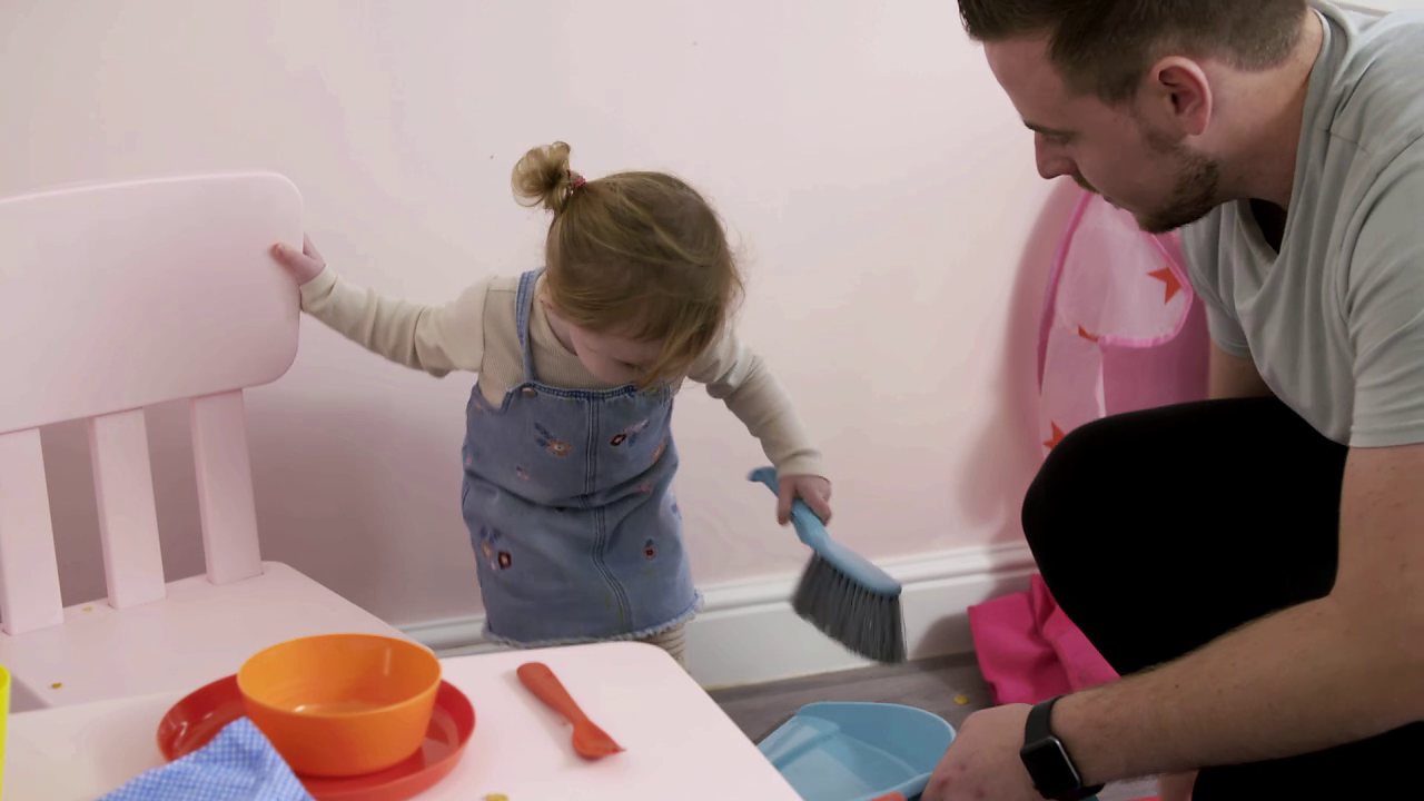 Little girl holding on to chair and crouching down to use a dustpan and brush