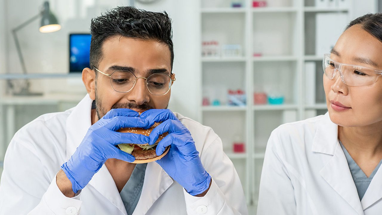A man in a white lab coat wearing blue gloves, eating a burger . A woman wearing a lab coat and goggles is watching him.