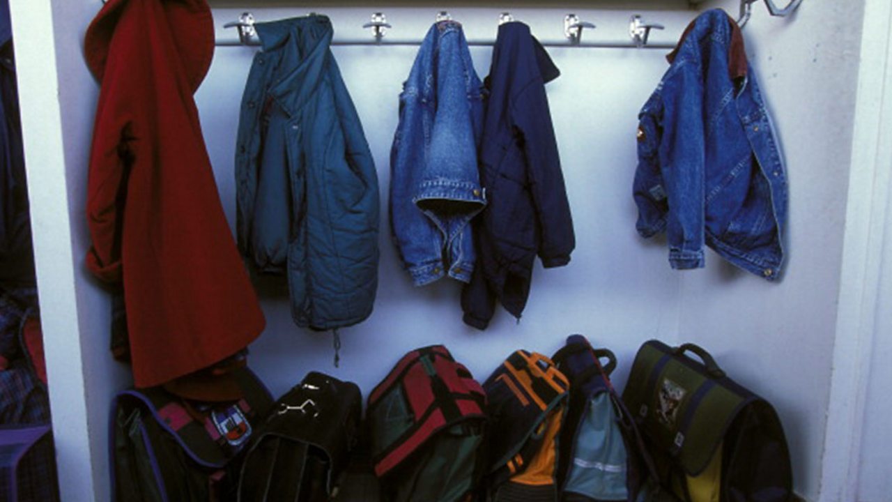 A collection of five coat and jackets hanging on a coat rack. Six school bags are neatly stored beneath the coat rack.