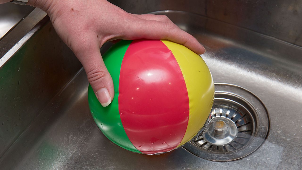 A hand holding a small inflatable ball in a sink filled with warm water. The ball is hard.