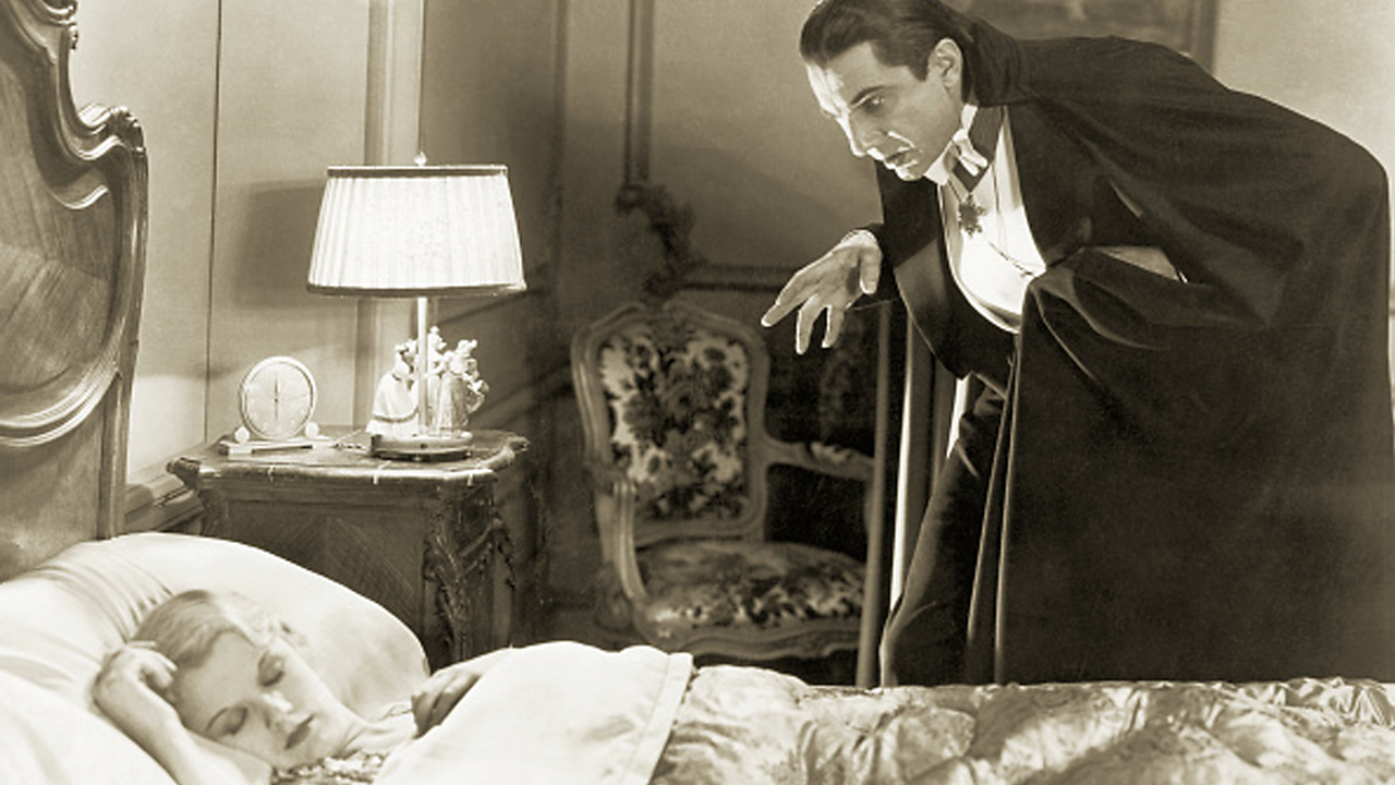 A man with dark hair and a black and white suit with a black cape leans over a woman asleep in bed.