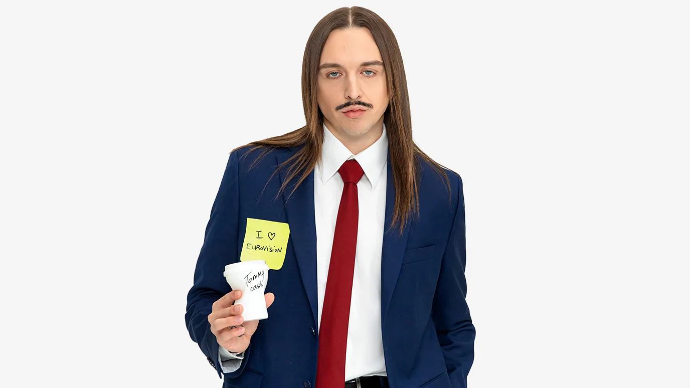Tommy Cash wearing a suit and holding a post-it note which reads "I he...