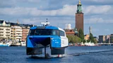 A blue-and-white electric hydrofoil ferry is lifted clear of the water as it cruises by a city waterfront (Credit: Candela)