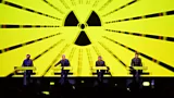 The four members of Kraftwerk performing on stage with a radioactive symbol against a bright neon yellow screen behind them (Credit: Getty Images)
