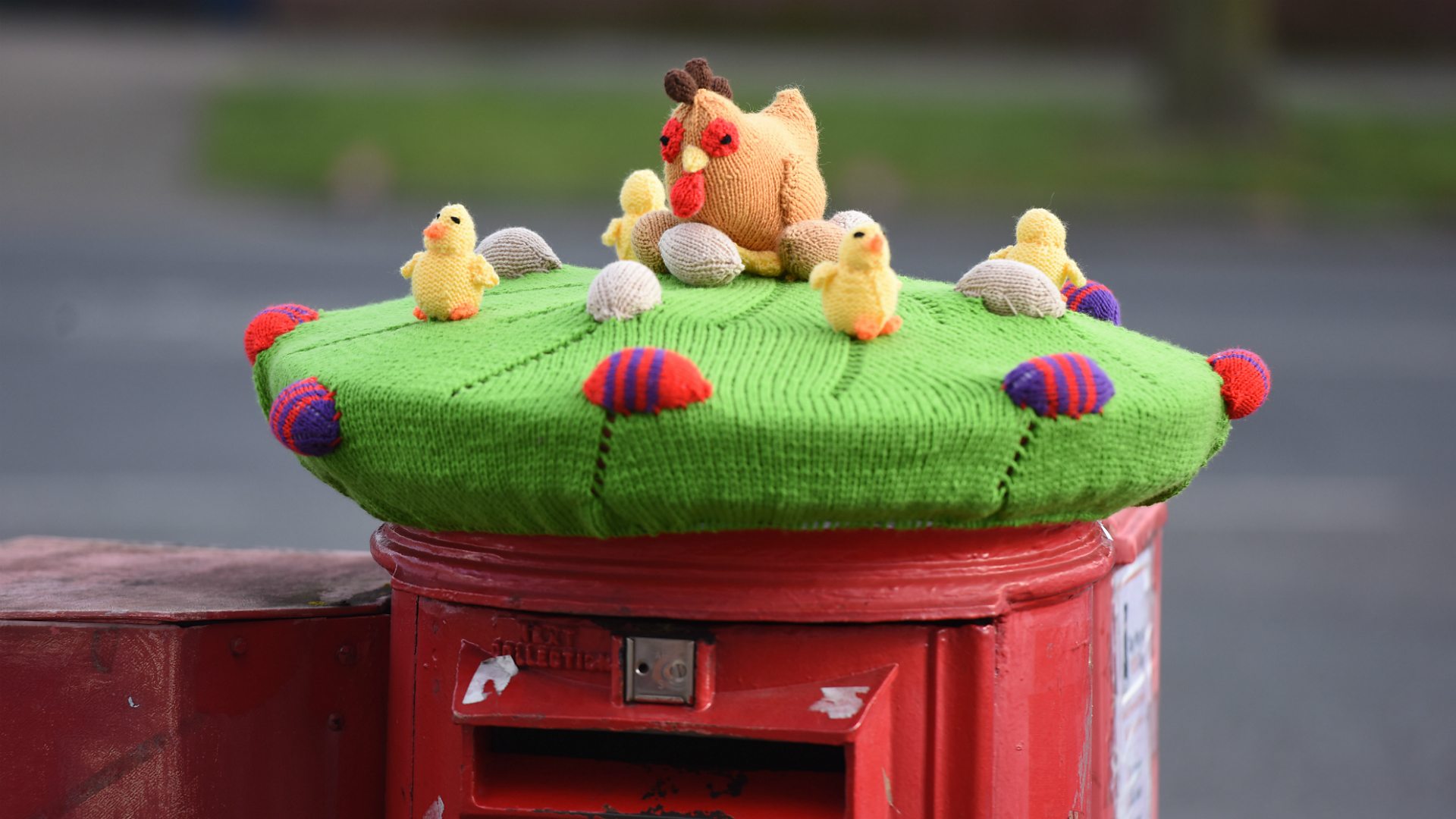 A knitted Easter postbox topper that shows a chicken, chicks and eggs frolicking on some grass