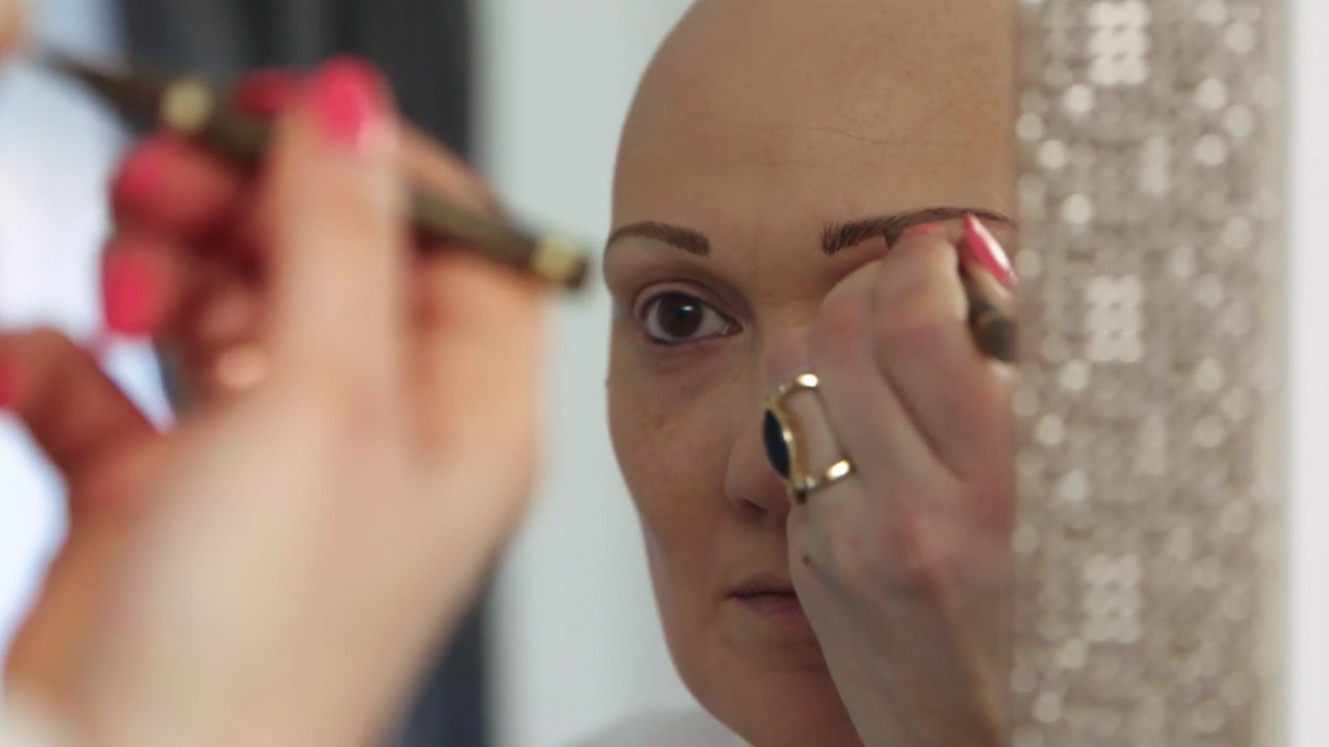 Carly, not wearing a wig, drawing on her eyebrows with an eyebrow pencil in the mirror