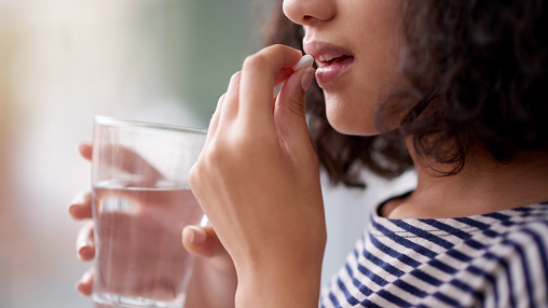 A woman holding a glass of water, putting a pill in her mouth