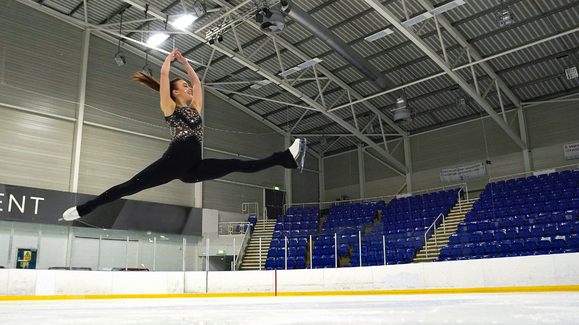 A photo of Emily doing a split jump on the ice, with her hands above her head.