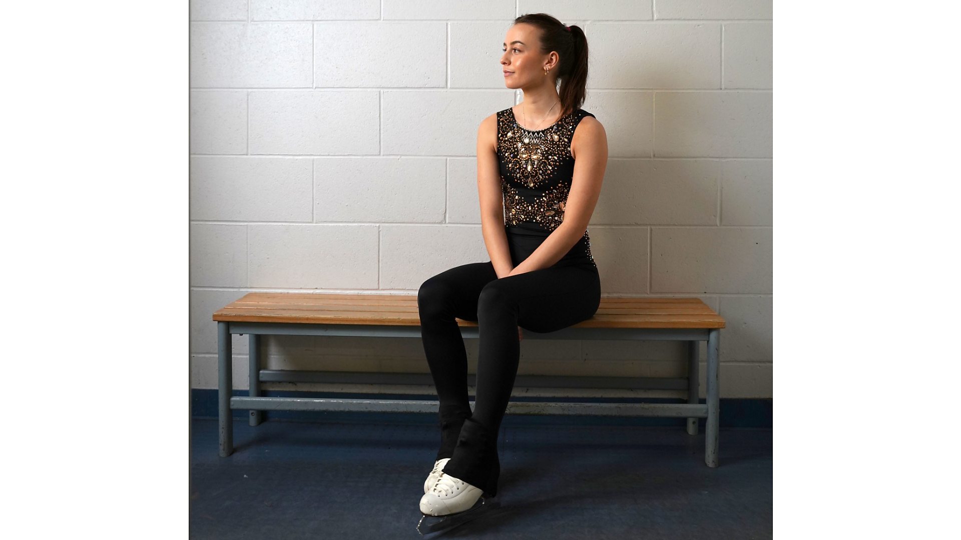 Emily sat in the changing rooms of an ice rink, wearing her ice skates, looking to side.