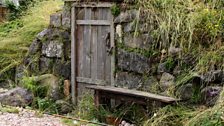 A gravel path leads to the door of a humble house, which nestles into an earth mound.